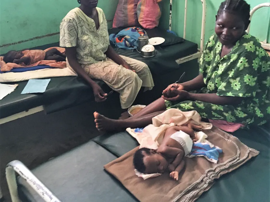 Two mothers sit on beds in the maternity ward, one with her baby sleeping on a blanket beside her